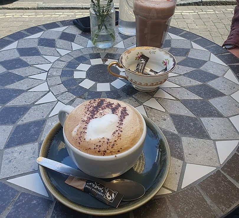 A cappuccino and an iced drink on a grey-and-white mosaic table, with pink carnations in a jar